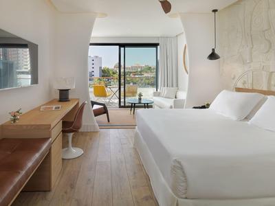 Moderní hotelový pokoj s velkou postelí, dřevěným nábytkem a balkonem s výhledem na město.