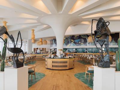 Moderní hotelová hala s bílým stropním designem, dřevěnou podlahou, sochami a kaktusy.