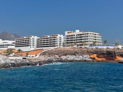 Velké bílé hotelové budovy na skalnatém pobřeží s klidným modrým mořem a jasnou oblohou.