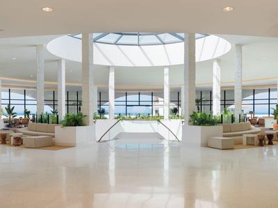 Světlá hotelová hala s kruhovým střešním oknem, béžovými pohovkami a rostlinami za přirozeného světla.