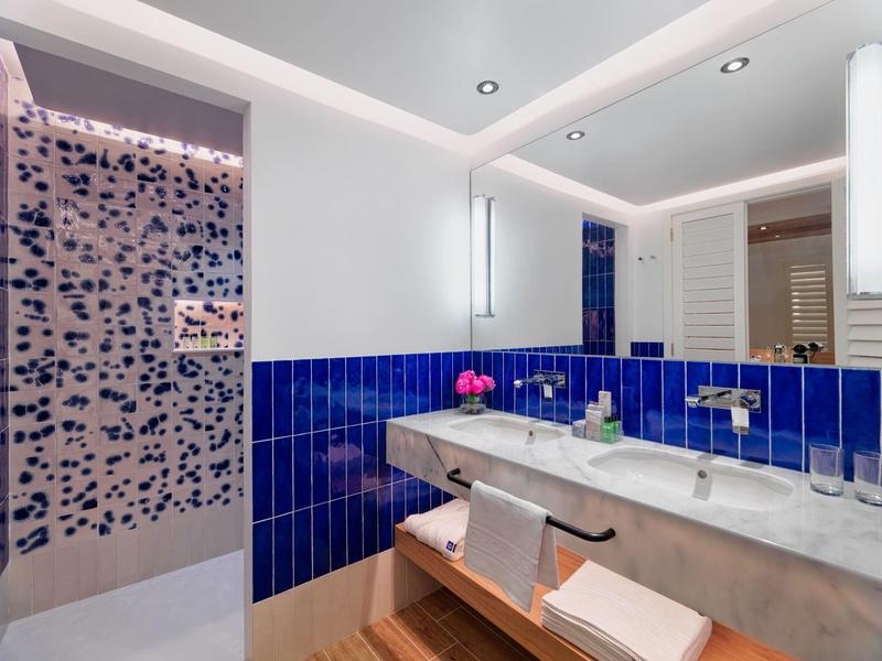 Moderní koupelna s modrými dlaždicemi, dvojitým umyvadlem, velkým zrcadlem a skleněnými dveřmi sprchy.