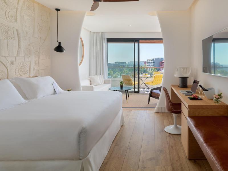 Moderní hotelový pokoj s velkou bílou postelí, dřevěnou podlahou, stolem a balkonem s výhledem.