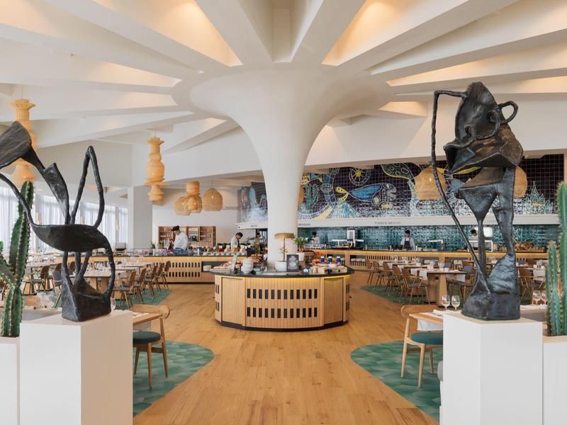 Moderní hotelová hala s bílým stropním designem, dřevěnou podlahou, sochami a kaktusy.