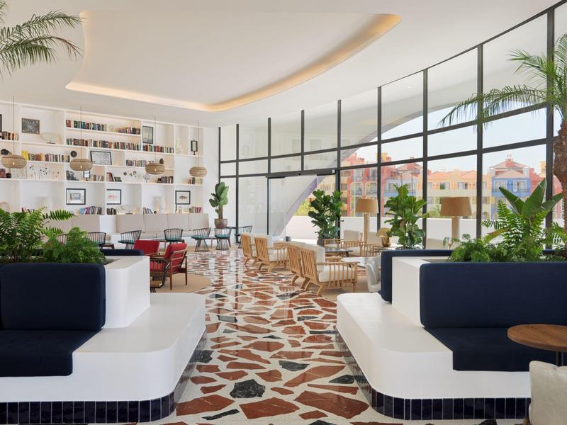 Světlý hotelový salonek s moderními bílými a námořnicky modrými pohovkami, rostlinami a velkými zakřivenými okny od podlahy ke stropu.