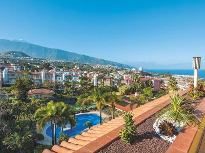 Výhled z terasy s rostlinami na město s bazénem a horami v pozadí.