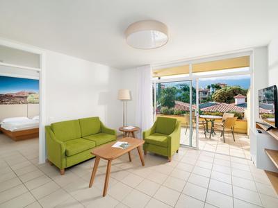 Jasný obývací pokoj se zelenou pohovkou, křeslem, TV a přístupem na balkon se stolem a židlemi.