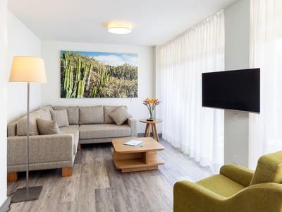 Moderně zařízený obývací pokoj s pohovkou, televizí, křeslem a velkými okny se závěsy.