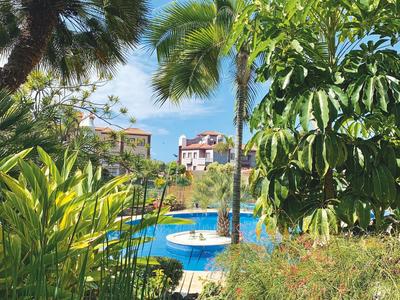 Modrý bazén s fontánou obklopený tropickými palmami a rostlinami pod modrou oblohou.