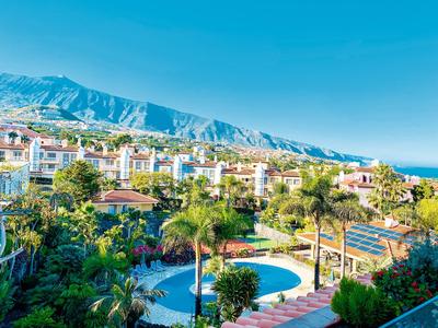 Pohled na hotelový resort s bazénem, palmami a horami v pozadí za jasné oblohy.