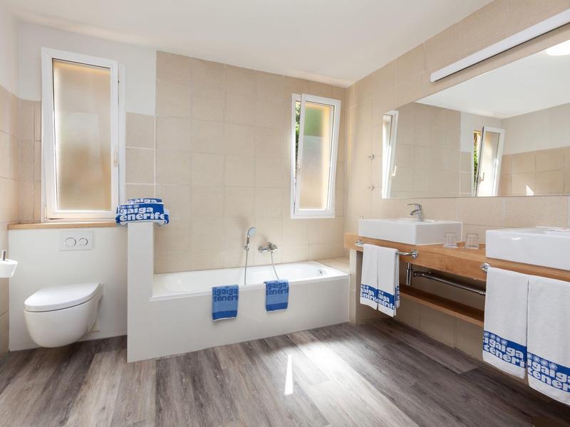 Moderní koupelna s vanou, toaletou, velkými zrcadly a dřevěnou deskou.