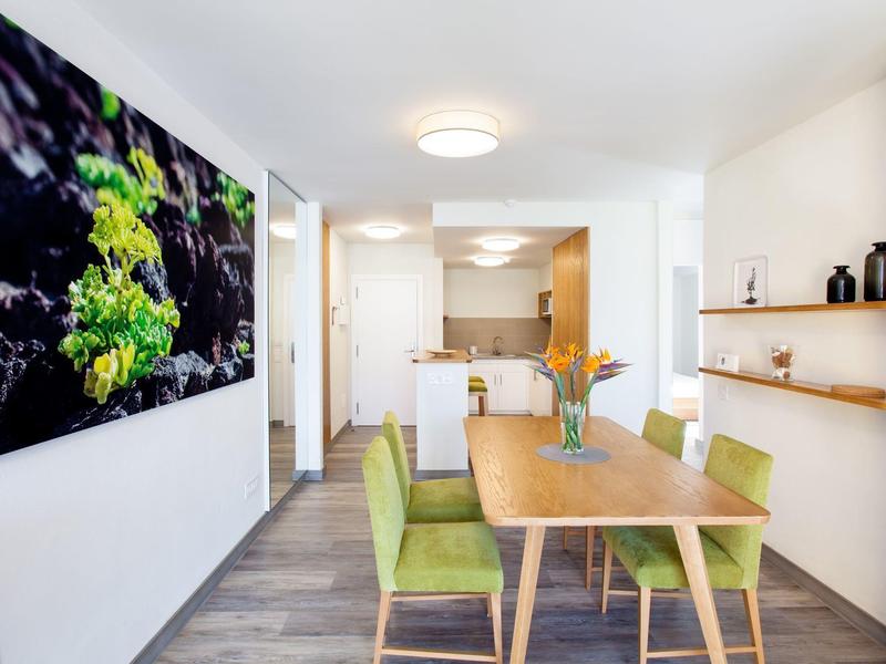 Moderní jídelní kout se zelenými čalouněnými židlemi a velkým nástěnným uměním.