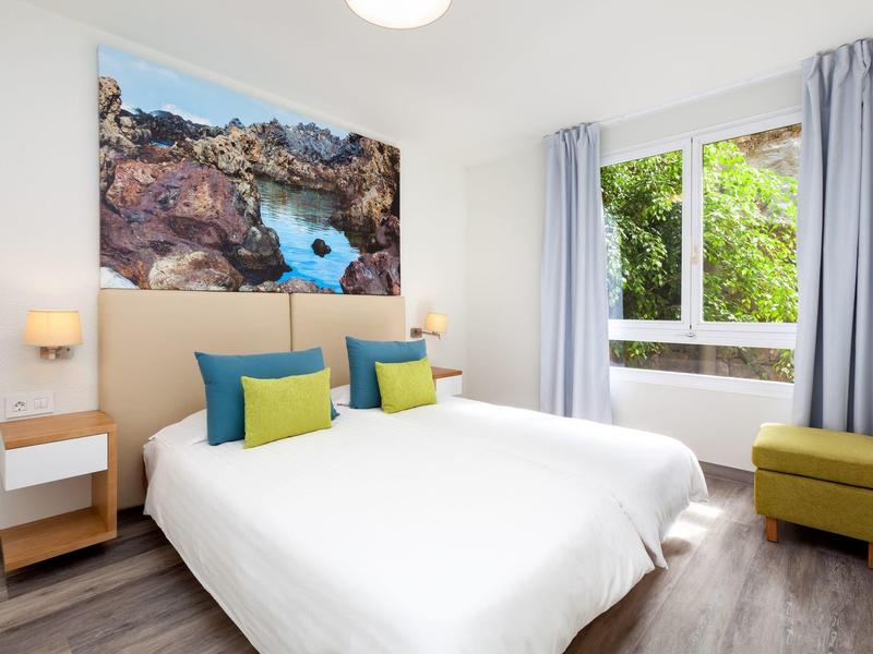 Moderní pokoj s dvěma jednolůžky, barevnými polštáři a velkým obrazem nad čelem postele.