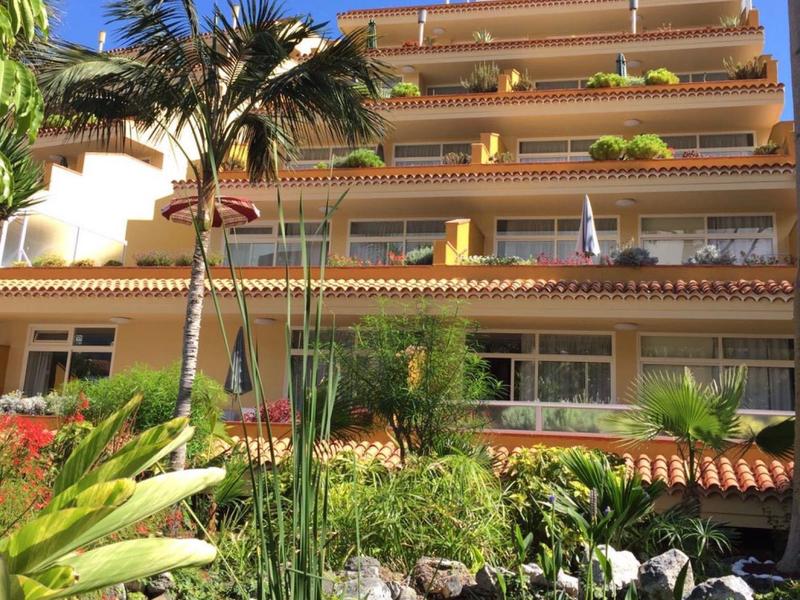 Vícepodlažní hotel s balkony, palmami a bujnou zahradou pod jasně modrou oblohou.