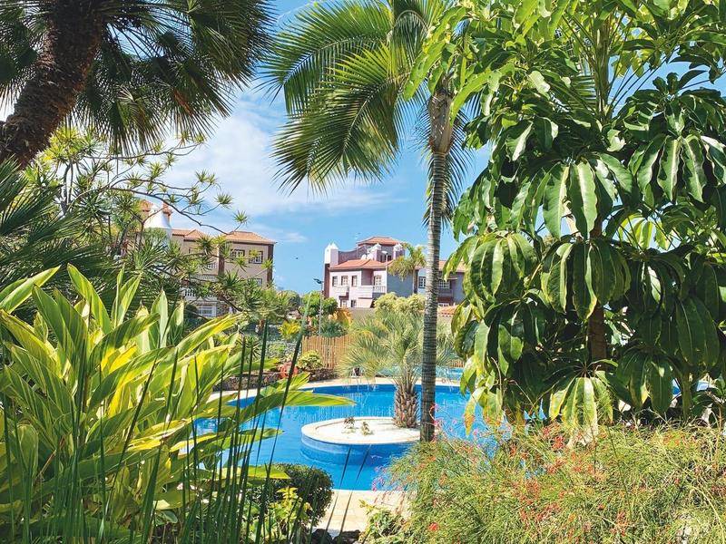 Pohled na modrý bazén s vodními nádržemi obklopený zelenými tropickými rostlinami a palmami pod jasnou oblohou.