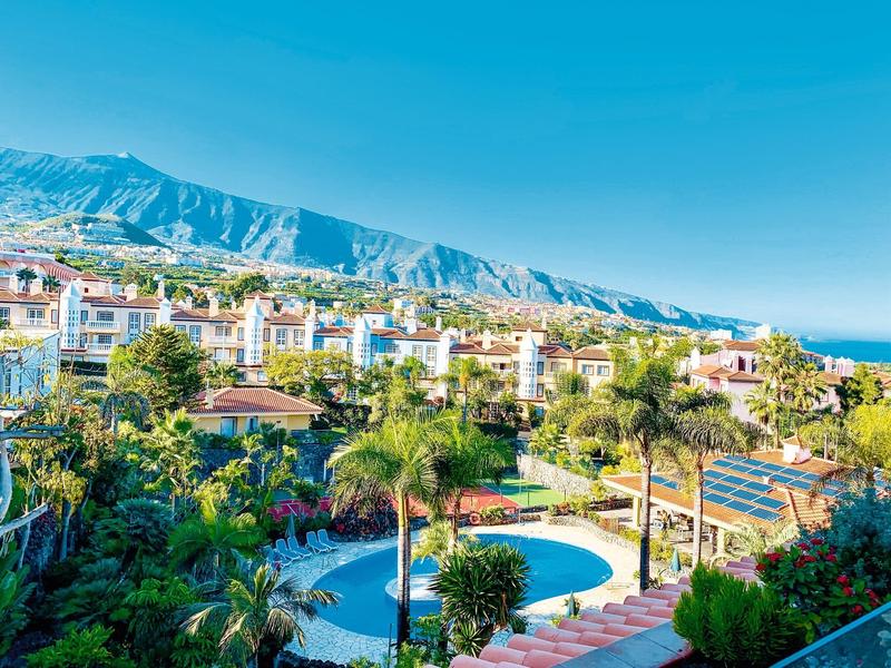 Rekreační resort s několika bazény, palmami a horami v pozadí pod jasnou oblohou.