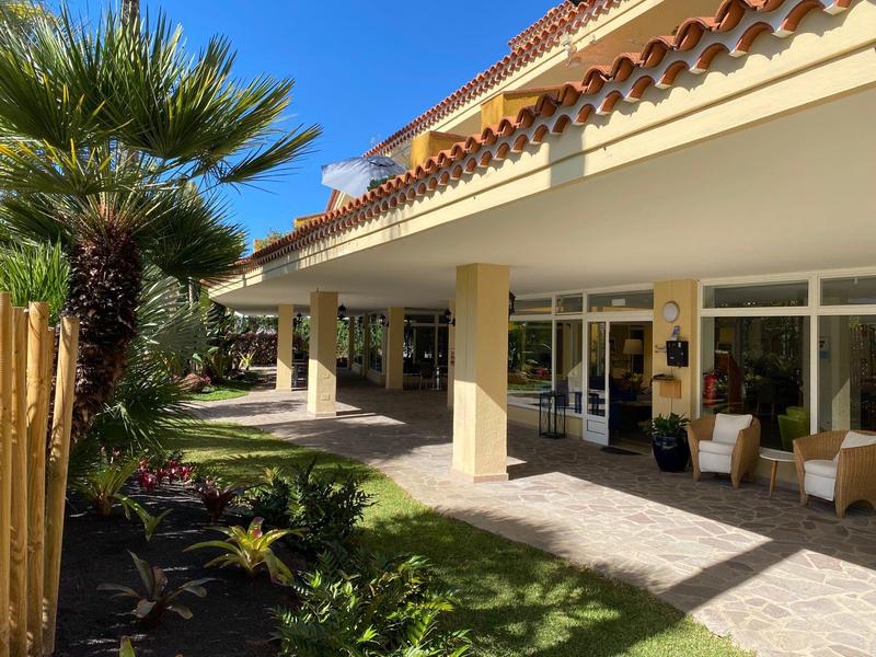 Zastíněná terasa hotelu s rostlinami a židlemi za slunečného dne.