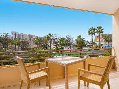 Balcone con due sedie e un tavolo che si affaccia su una piscina e palme sotto un cielo sereno.