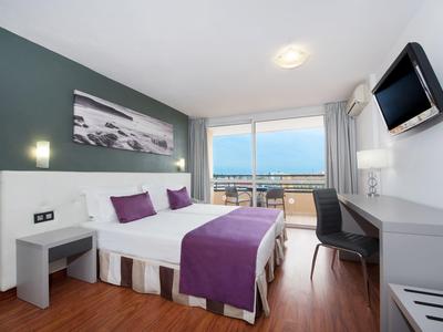 Camera d'hotel moderna con letto matrimoniale, accenti viola e balcone con vista mare.