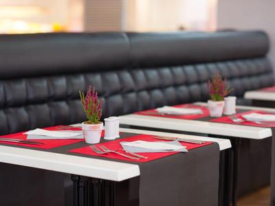 Tavoli con tovagliette rosse, piatti bianchi e piccole piante in un ristorante con panche nere.