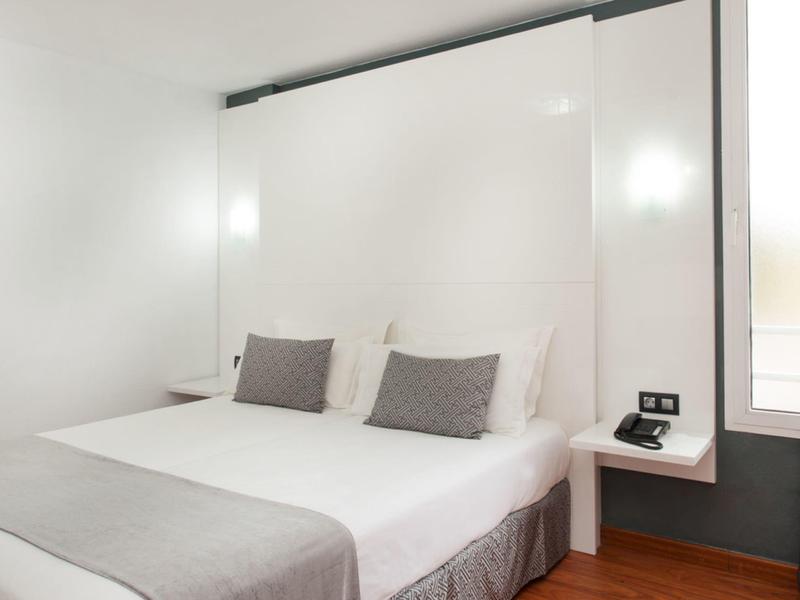 Camera d'hotel moderna con letto matrimoniale, cuscini grigi e illuminazione minimalista.