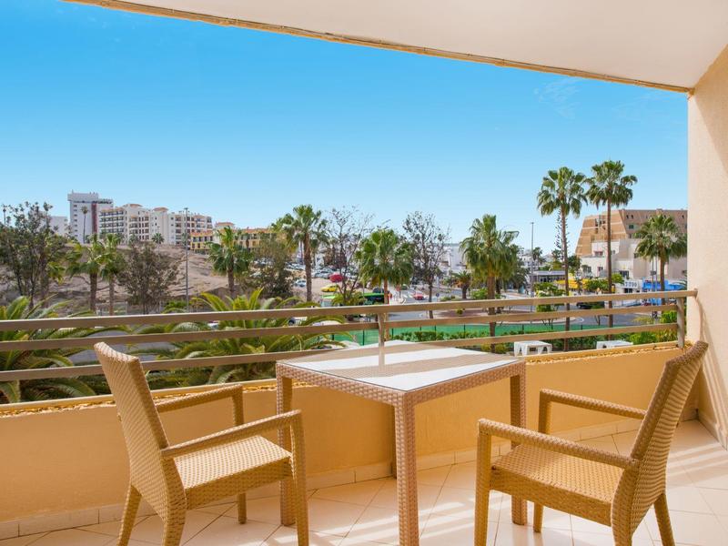 Balcone con due sedie e un tavolo che si affaccia su una piscina e palme sotto un cielo sereno.