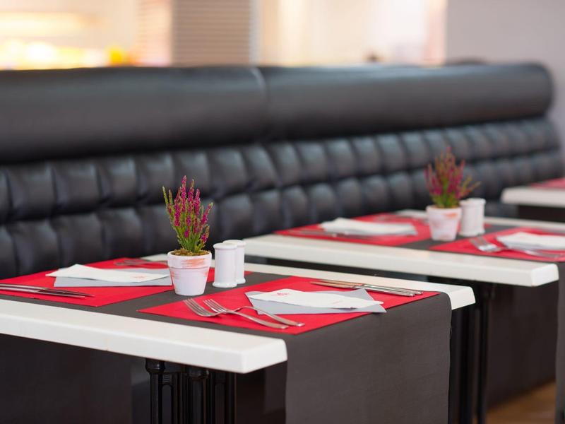 Tavoli con tovagliette rosse, piatti bianchi e piccole piante in un ristorante con panche nere.