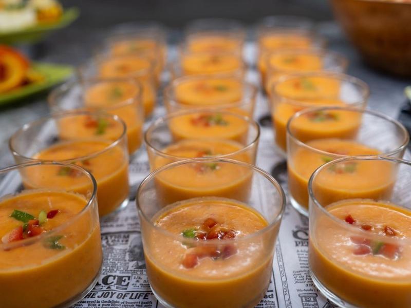 Rij van kleine glazen met oranje soep, gegarneerd met kruiden en spek.