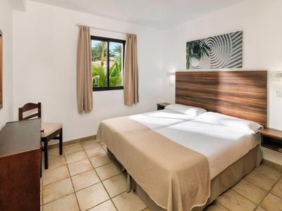 Ein schlichtes Zweibettzimmer mit hellbraunem Fußboden, weißer Wand und Fenster mit Vorhängen.