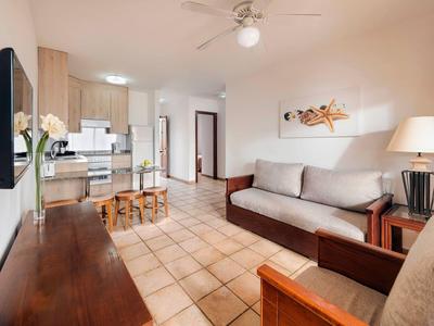 Helles Wohnzimmer mit beiger Couch, Holzmöbeln, Fliesenboden und offener Küche im Hintergrund.