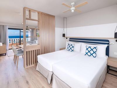 Helles Hotelzimmer mit zwei Betten, Holzlamellen-Trennwand und Blick auf das Meer.