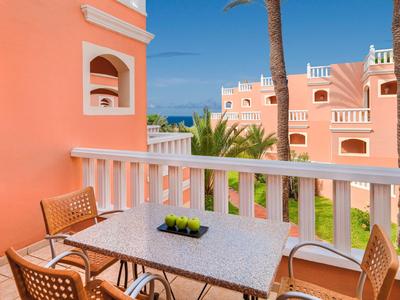 Balkon mit Marmor-Tisch, zwei Stühlen, Palmen und rosa Gebäuden unter blauem Himmel.