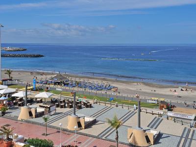 Strand mit Sand, Meer und blauem Himmel, Strandliegen und Menschen am Ufer, Promenade mit Palmen.