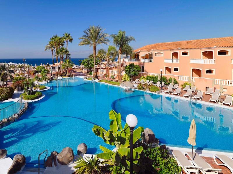 Velký modrý bazén obklopený palmami a okrovou budovou pod jasnou oblohou.