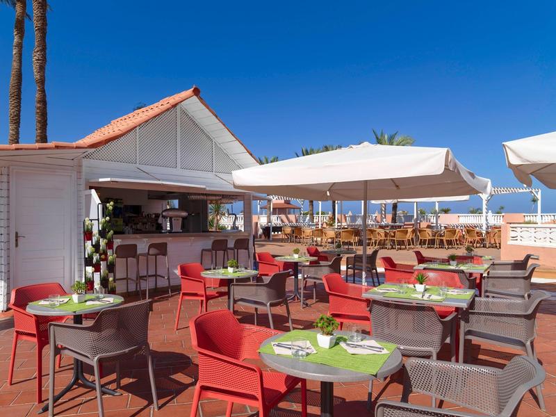 Venkovní terasa s červenými stoly a židlemi pod modrou oblohou vedle baru.