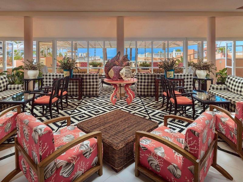 Barevný hotelový salón s květinovými křesly, geometrickou podlahou a výhledem na moře velkými okny.