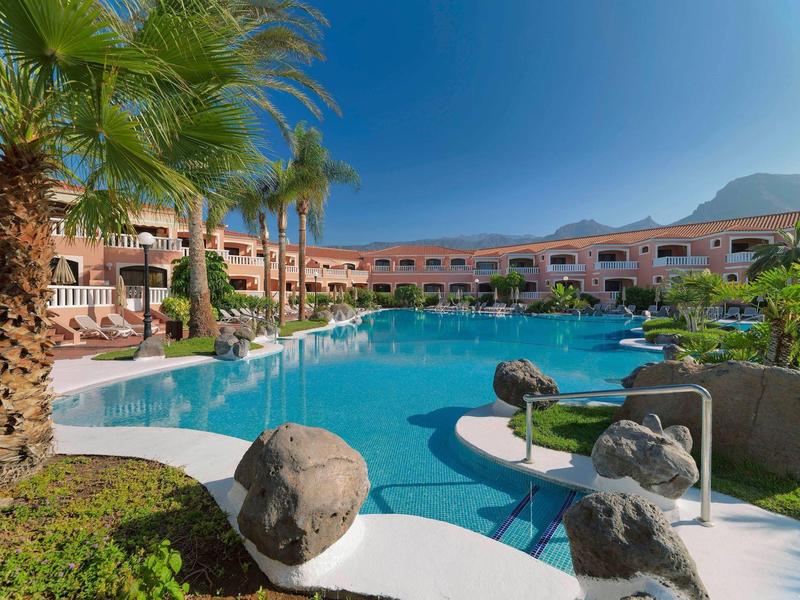 Velký bazén obklopený palmami a budovami hotelu pod modrou oblohou.