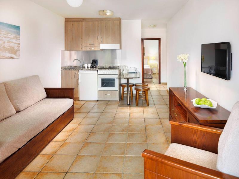 Obývací pokoj s vybavenou kuchyní, pohovkou, jídelním stolem, křeslem a televizí na stěně.