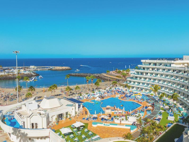 Resort con grande piscina, palme e vista mare in una giornata soleggiata.