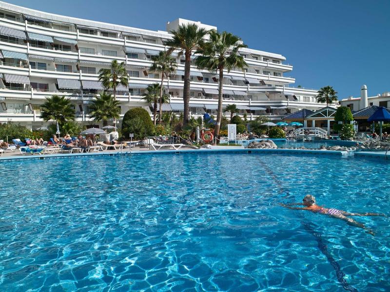 Large outdoor pool with clear blue water in front of a multi-story hotel building and palm trees.