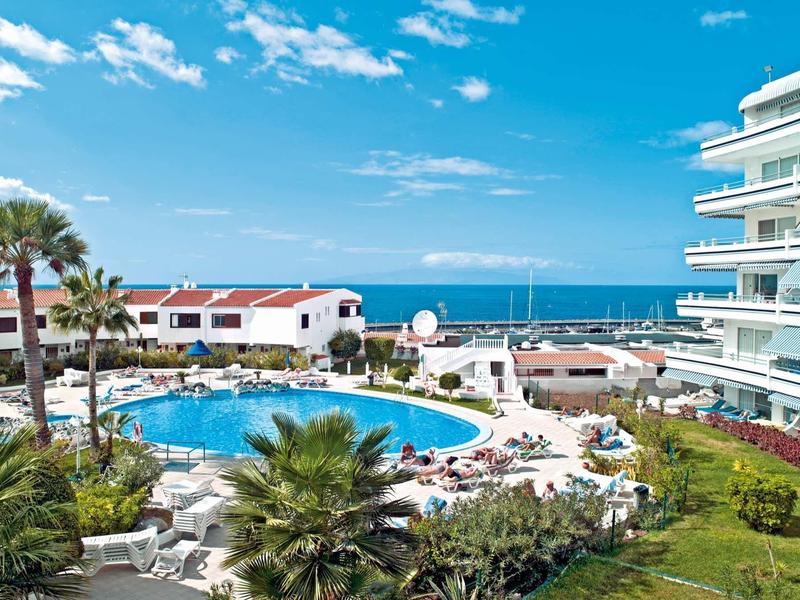 Hotel with round pool, sun loungers, palm trees, and sea view in the background.