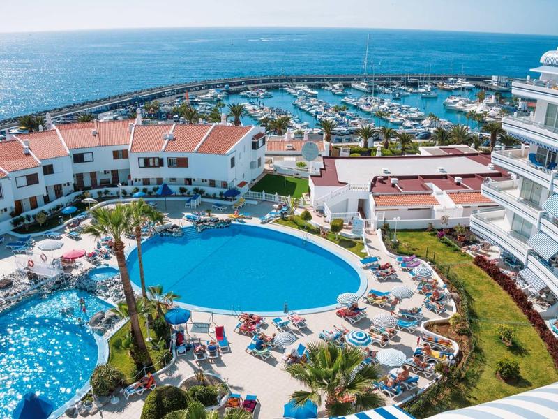 View of hotel pool, sun loungers, and marina by the sea on a sunny day.