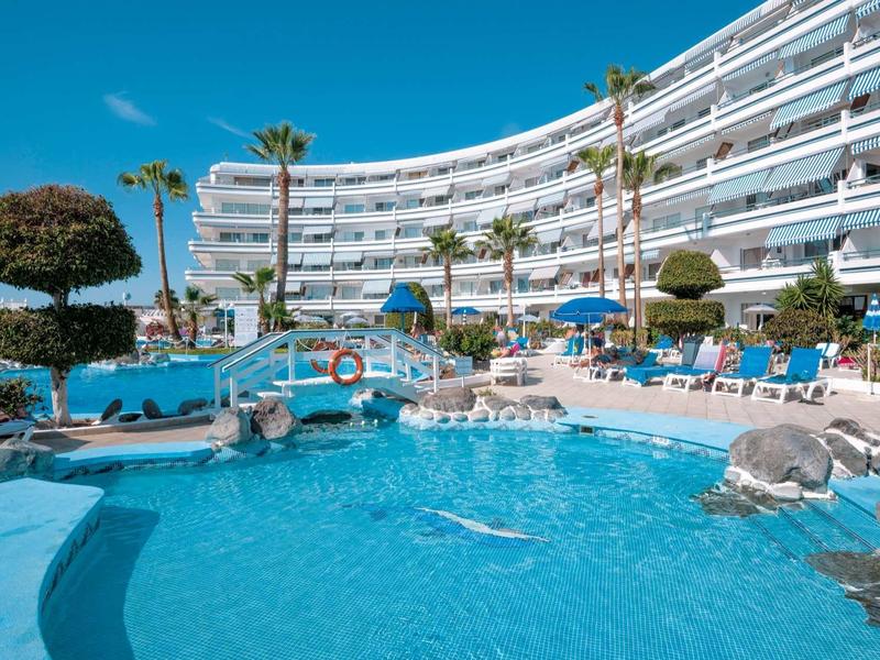 Hotel with large pool, sun loungers, and palm trees in front of a multi-story white building.