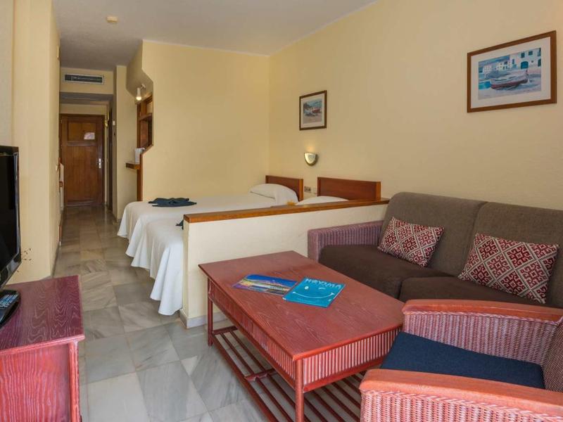 Hotel room with two beds, sofa, armchairs, and flat-screen TV in a bright space.