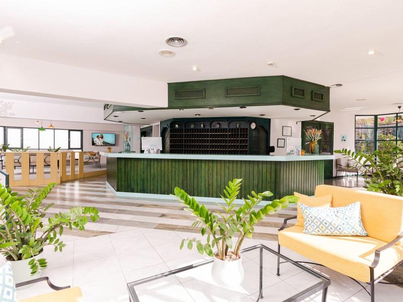 Jasny hol hotelowy z zielonym biurkiem recepcyjnym, żółtymi sofami i roślinami