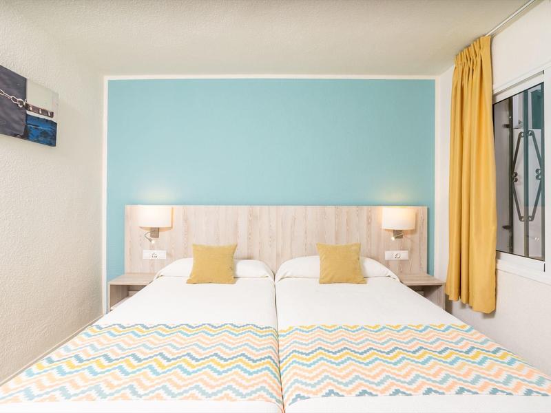 Dwa pojedyncze łóżka z kolorową pościelą na tle jasnoniebieskiej ściany i żółtych zasłon