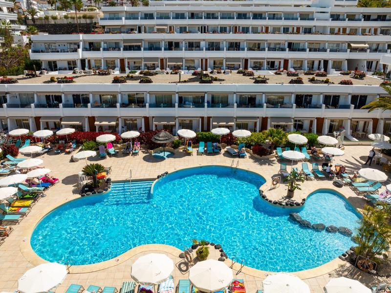 Duży hotelowy basen z parasolami i leżakami przed wielopiętrowym budynkiem hotelu.