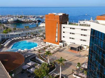 Hotel mit blauem Pool, weißen Liegestühlen, Palmen und Blick auf das blaue Meer im Hintergrund.