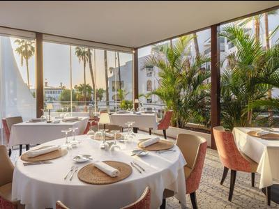 Elegantes Restaurant mit runden Tischen, weißen Tischdecken und großen Fenstern mit Palmenblick.