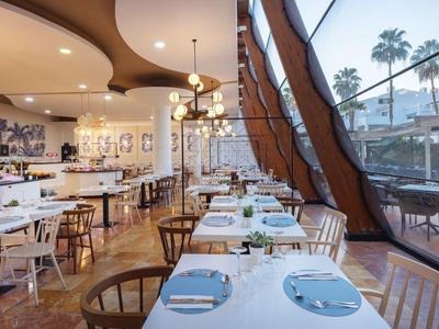 Modernes Restaurant mit Holzboden, weißen Tischen, Stühlen und großem Fenster mit Aussicht.