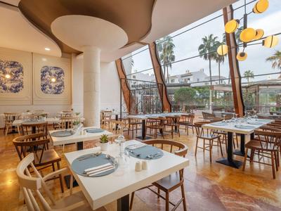 Zona ristorante luminosa arredata con mobili in legno e grandi finestre con vista sulle palme.